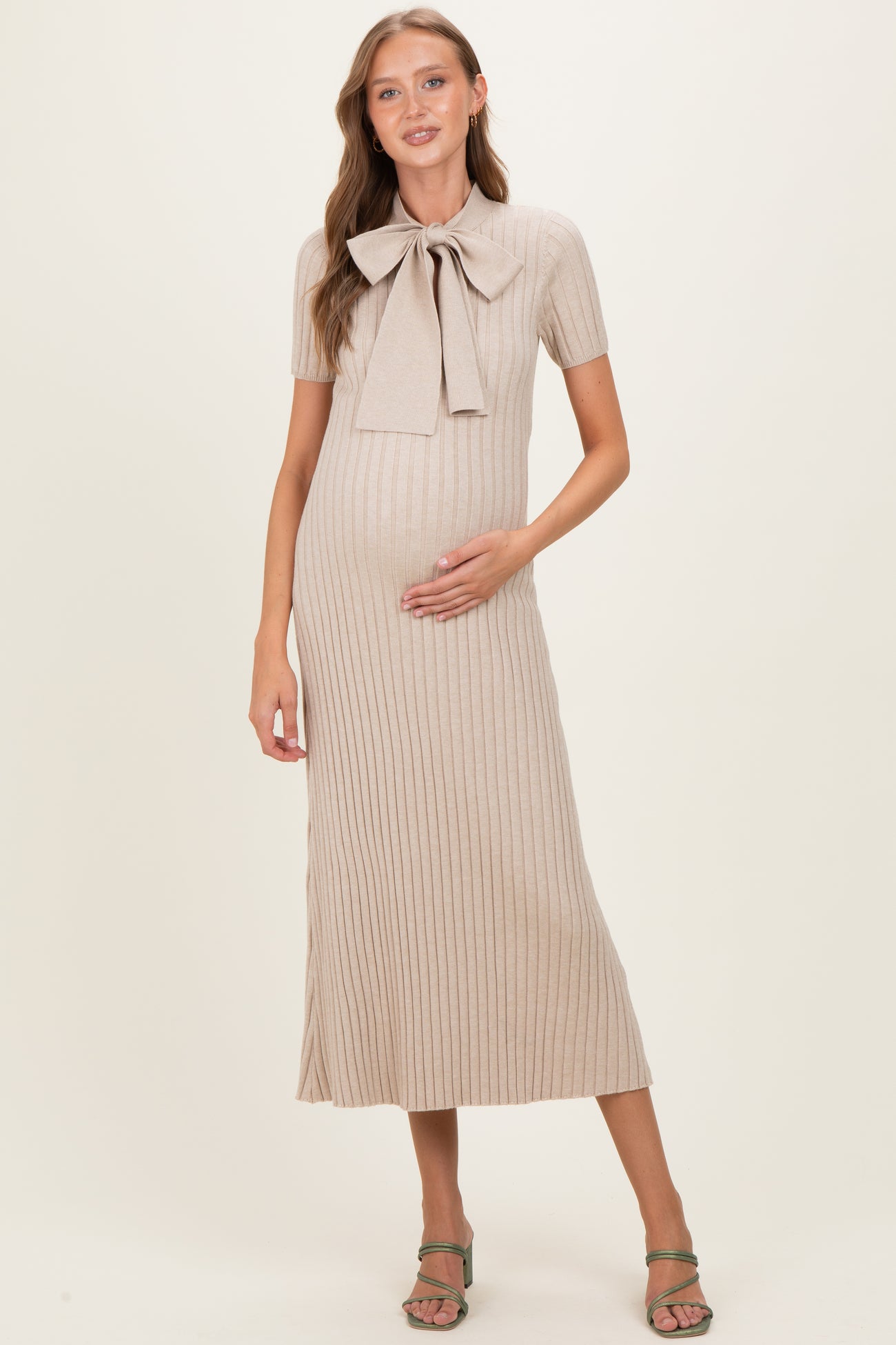 woman wearing a beige ribbed maternity dress with a bow tie detail and holding her baby bump