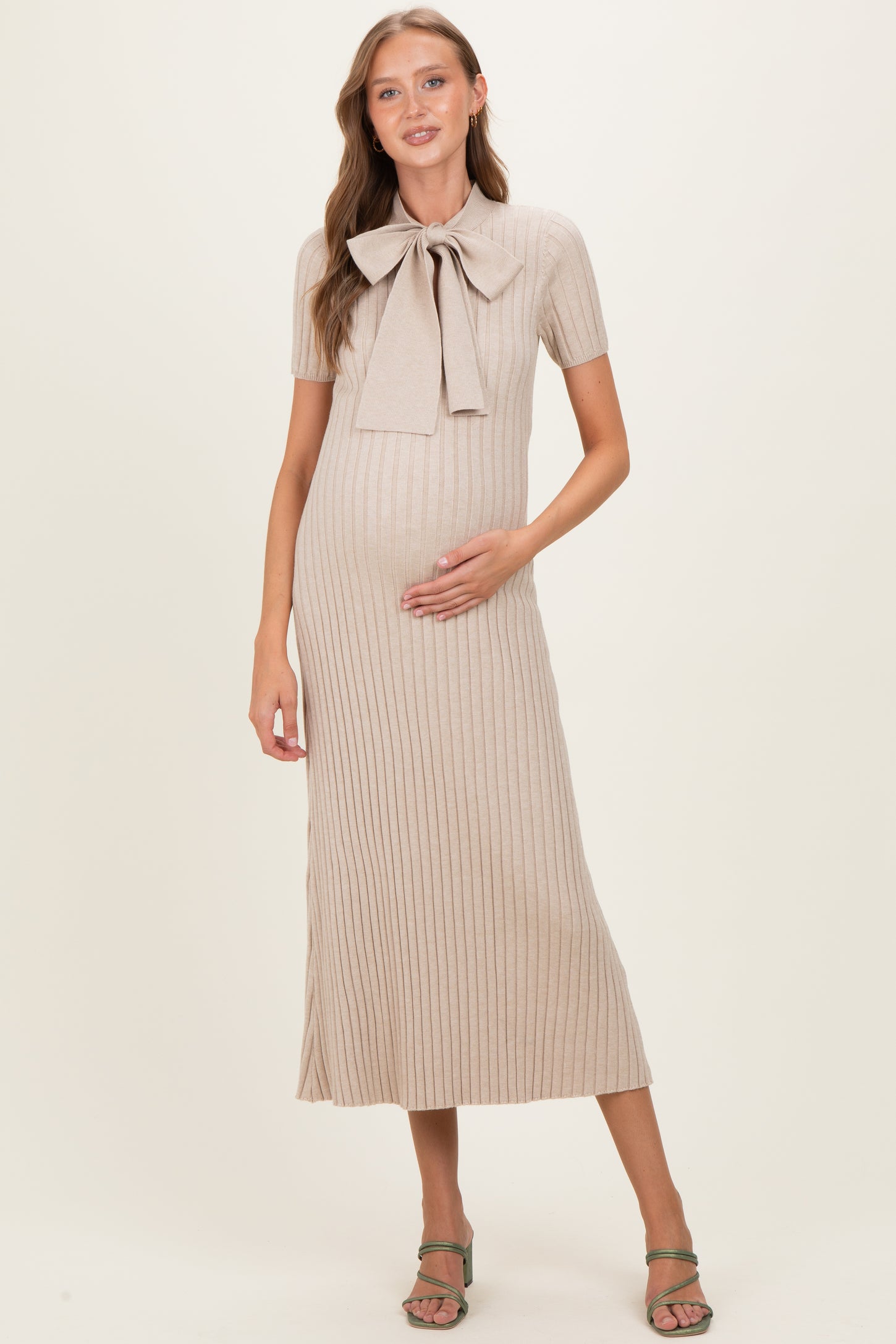 woman wearing a beige ribbed maternity dress with a bow tie detail and holding her baby bump