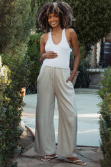 woman wearing a white maternity tank top and light beige wide-leg pants, holding her baby bump outdoors