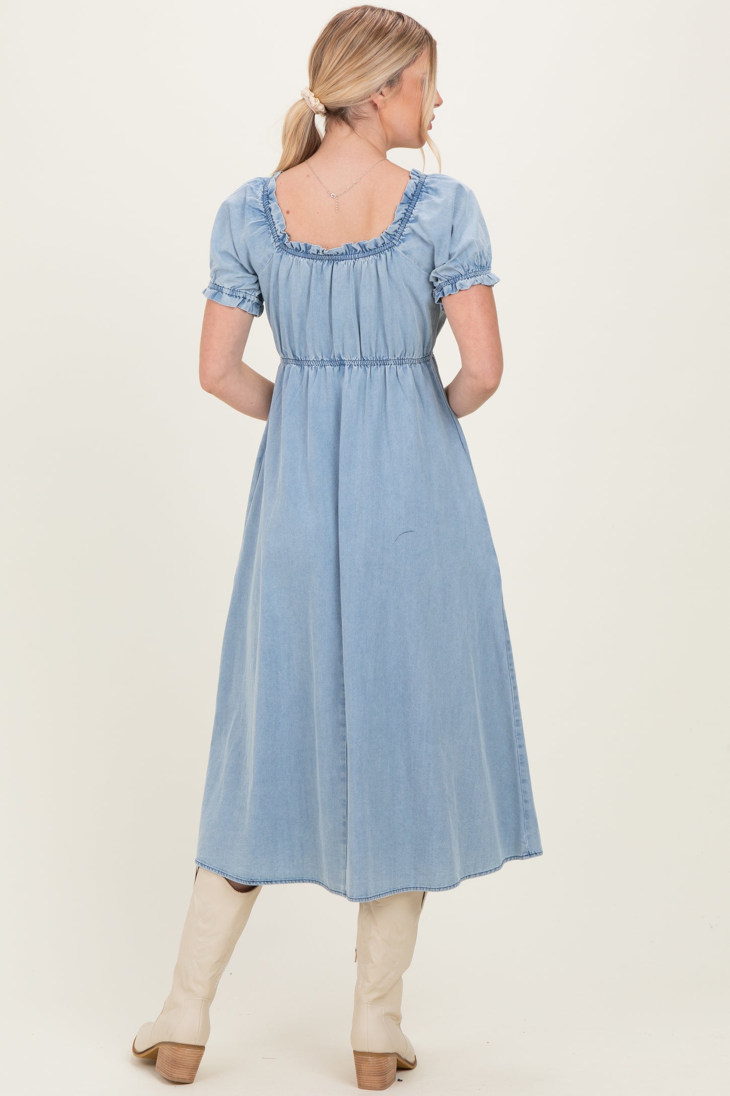 woman wearing a light blue denim maxi dress with puffy sleeves and a ruffled neckline, facing away from the camera