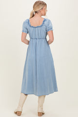 woman wearing a light blue denim maxi dress with puffy sleeves and a ruffled neckline, facing away from the camera