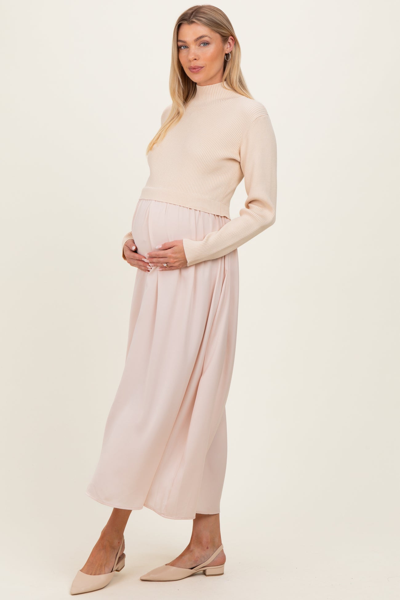 woman wearing a cream ribbed maternity sweater and a flowing blush pink maternity maxi skirt, holding her baby bump