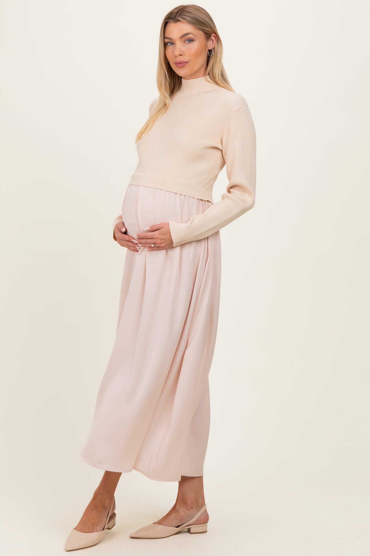 woman wearing a cream ribbed maternity sweater and a flowing blush pink maternity maxi skirt, holding her baby bump