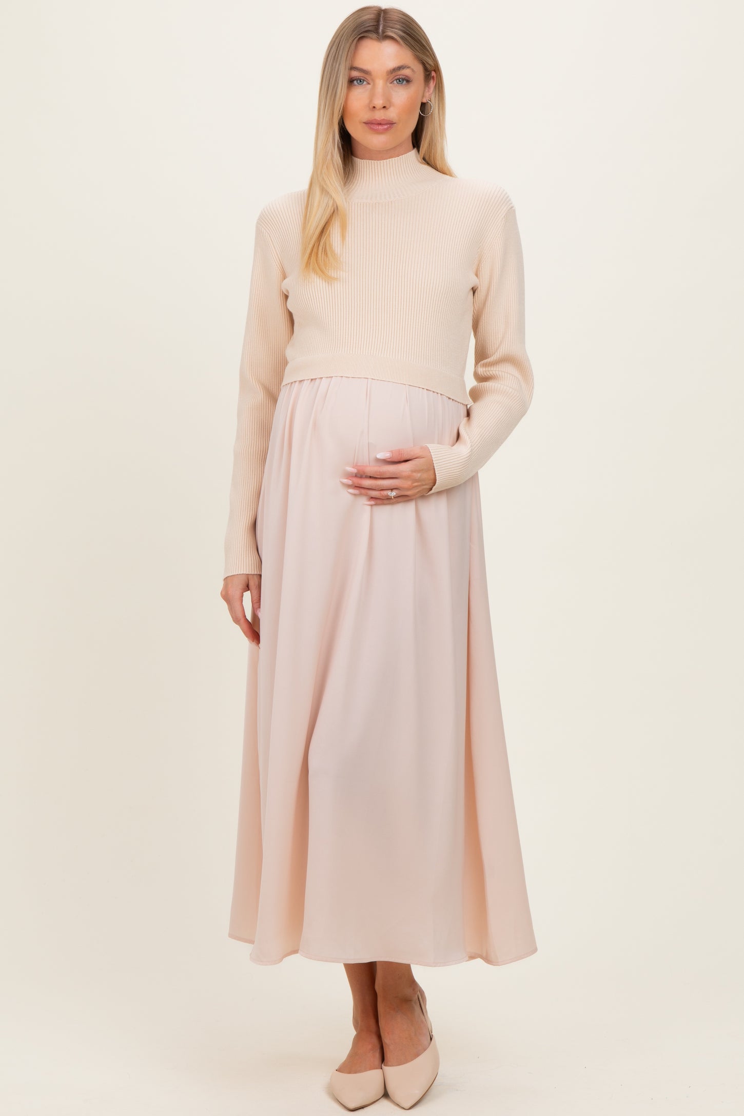 woman wearing a cream ribbed maternity sweater and a pale pink maxi skirt, gently holding her baby bump