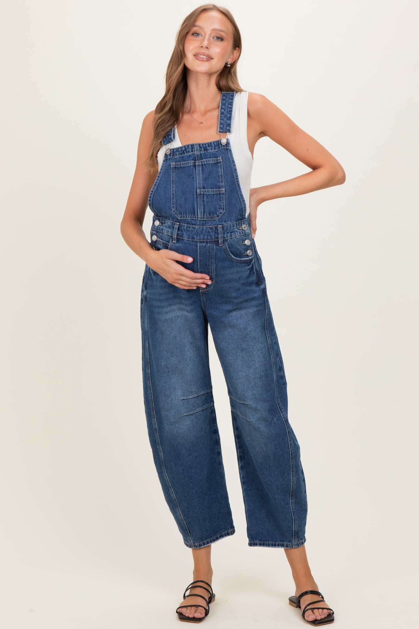 woman wearing a blue denim maternity overall with a white tank top, holding her baby bump and smiling