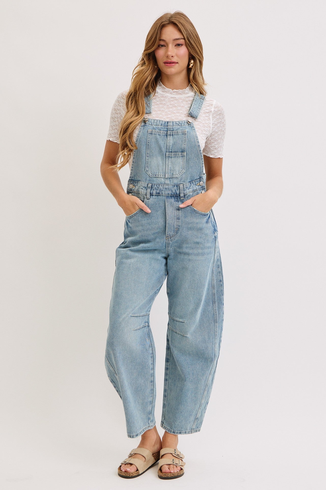 woman wearing light blue denim overalls with wide legs and a white short sleeve top, standing with hands in pockets