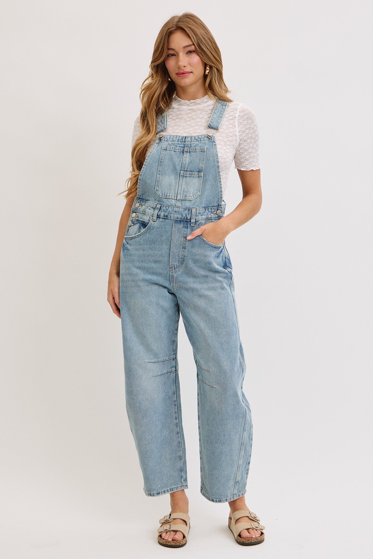 woman wearing light blue denim overalls with a white top, standing with hands in pockets on a neutral background