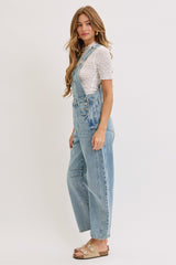 woman wearing light blue denim overalls over a white polka dot top, standing sideways with long hair and beige sandals