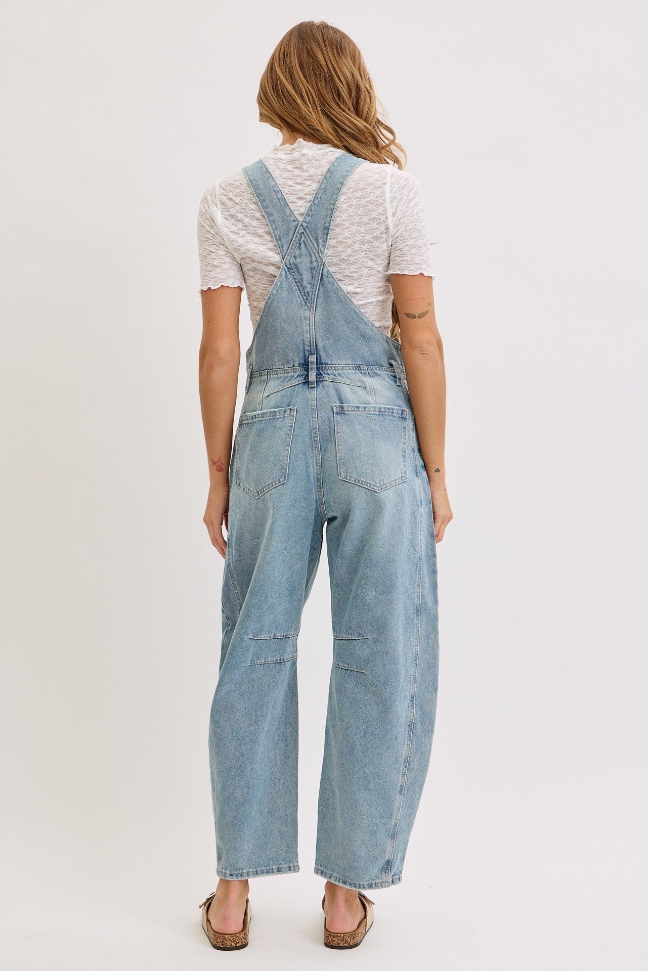 woman wearing a light blue denim overall with a loose fit and back bib detail, styled with a white top and sandals