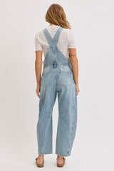 woman wearing a light blue denim overall with a loose fit and back bib detail, styled with a white top and sandals