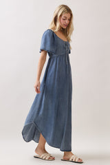 woman wearing a denim blue maxi dress with puff sleeves and a tie detail at the neckline