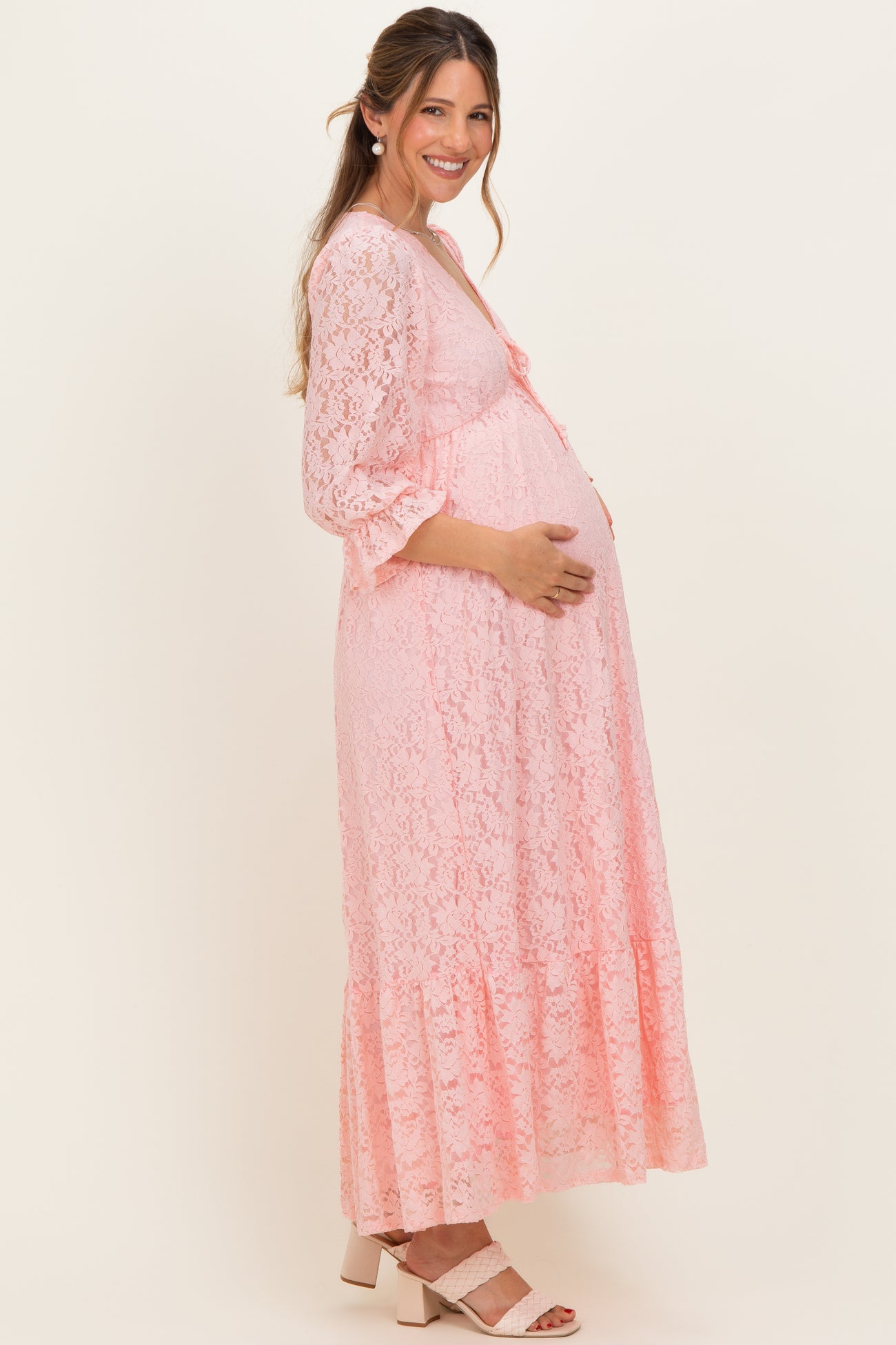 woman wearing a light pink lace maternity dress with three-quarter sleeves and a tiered hem, holding her baby bump