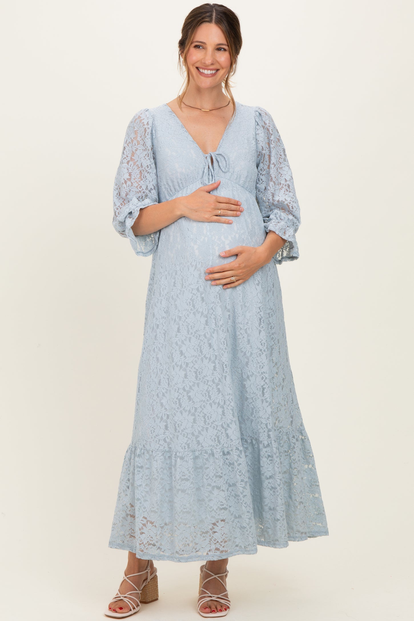 woman wearing a light blue lace maternity dress with a tie front and puff sleeves, cradling her baby bump