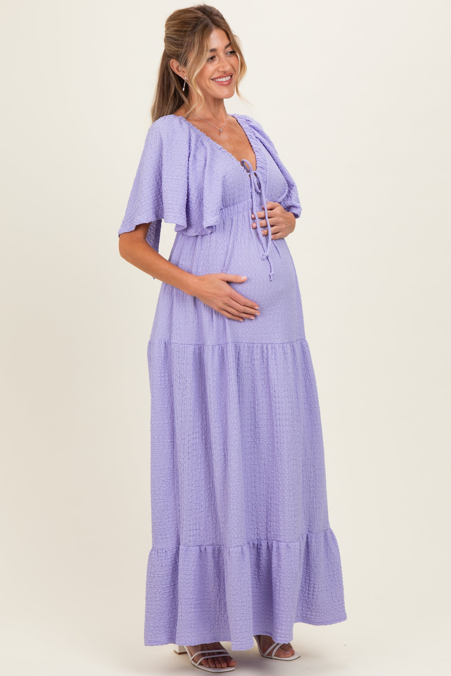 woman wearing a lavender maternity maxi dress with short sleeves and a tie-front detail, holding her baby bump