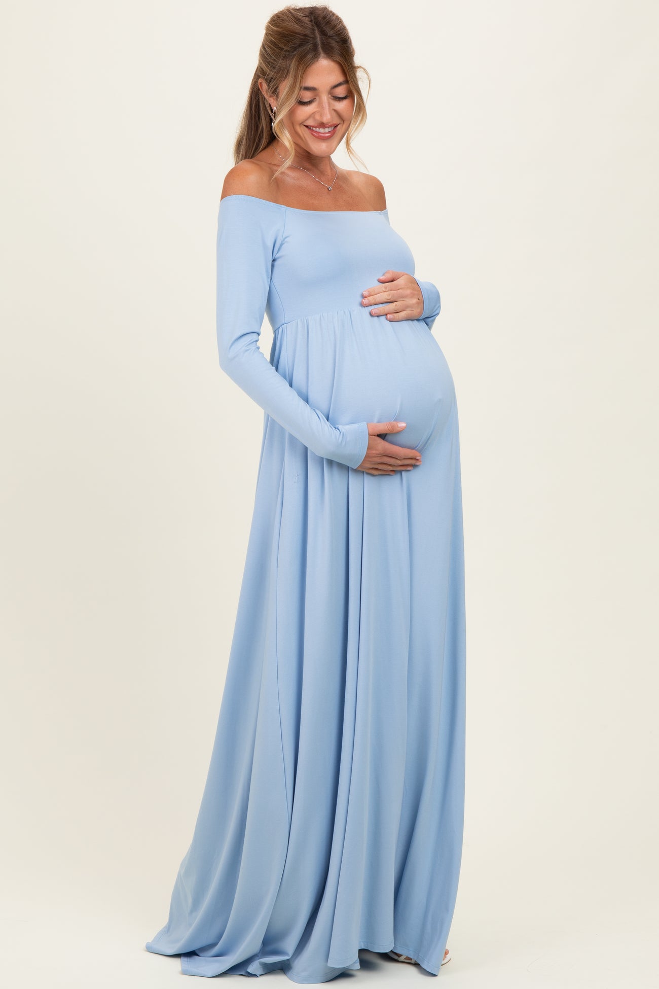 woman wearing a light blue off-shoulder maternity dress with a flowing skirt and holding her baby bump
