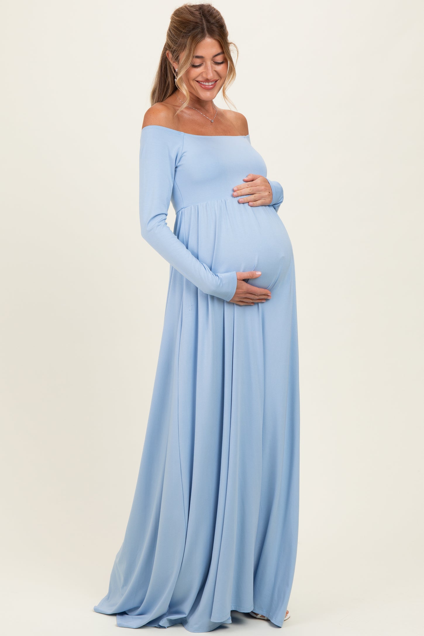 woman wearing a light blue off-shoulder maternity dress with a flowing skirt and holding her baby bump