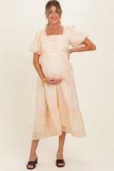 woman wearing a light peach and white gingham maternity dress with puff sleeves and a tiered hem, holding her baby bump