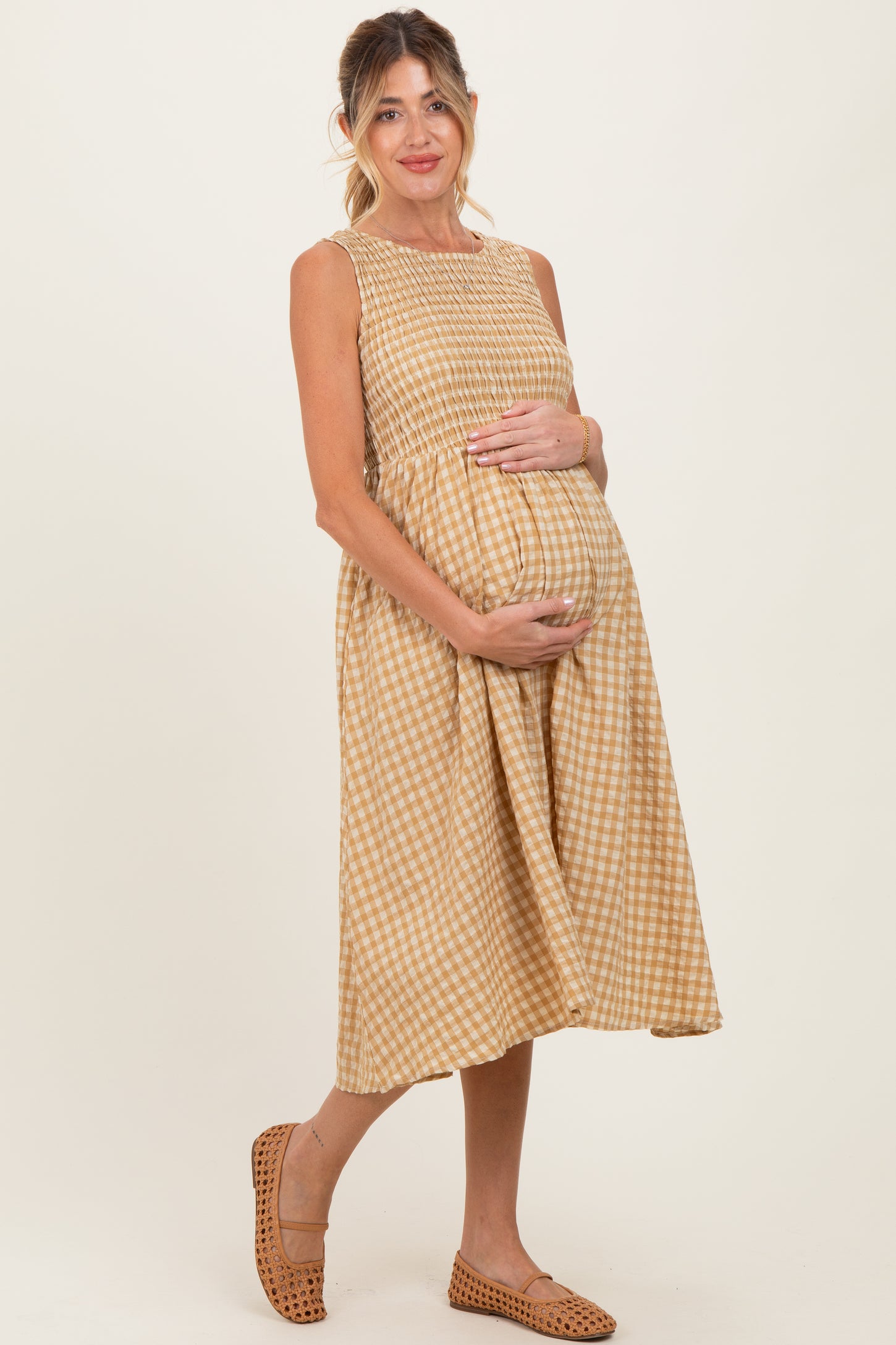 woman wearing a tan and cream gingham maternity dress and cradling her baby bump