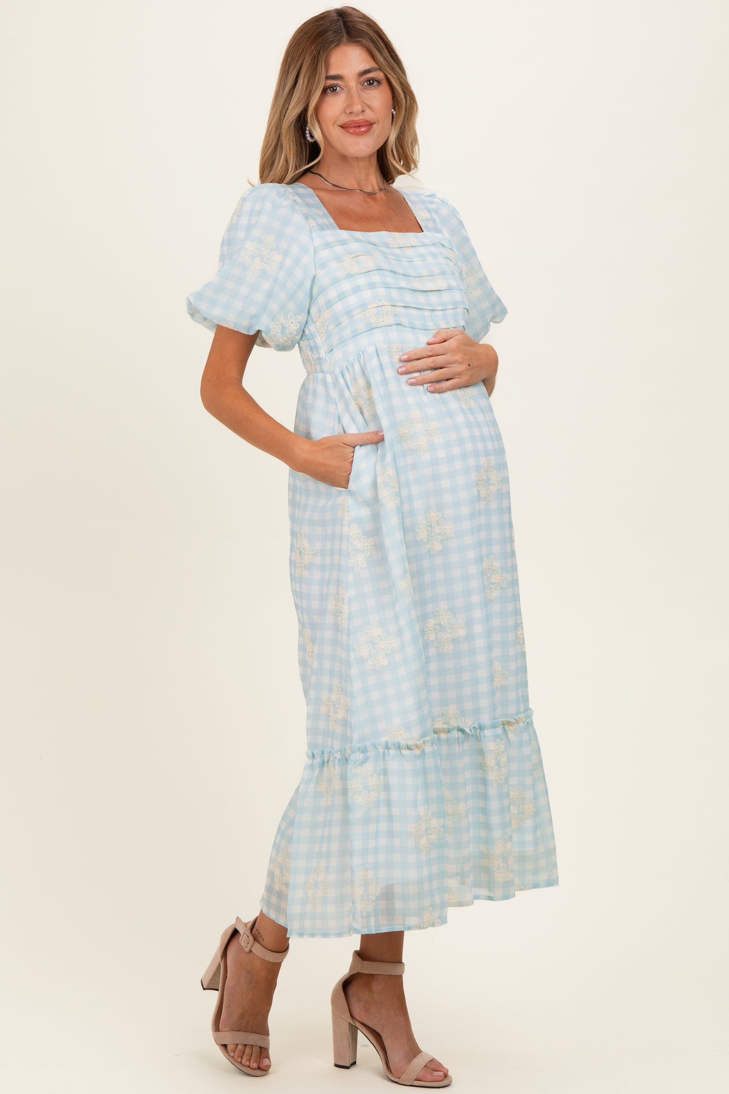 woman wearing a light blue gingham maternity dress with short puff sleeves and a ruffled hem, holding her baby bump