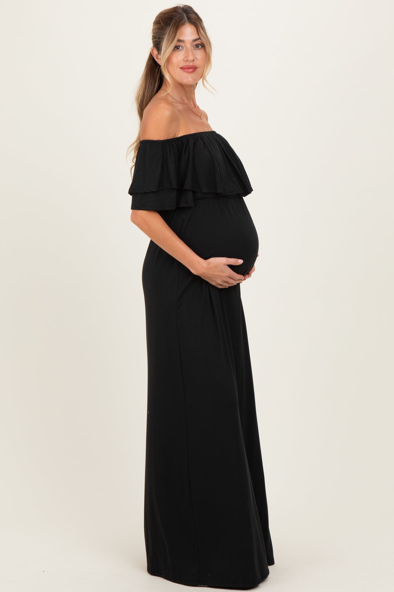 woman wearing a black off-the-shoulder maternity maxi dress, gently cradling her baby bump