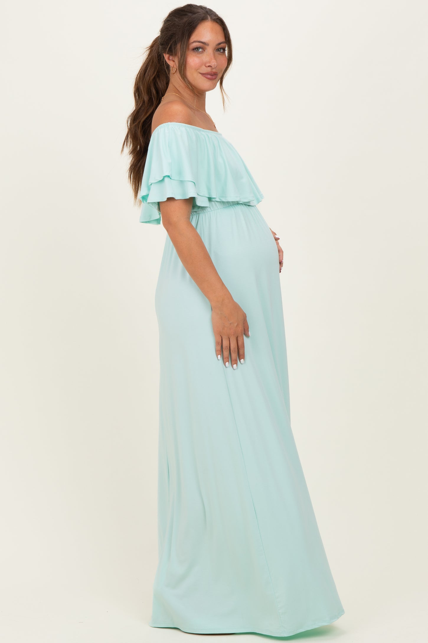 woman wearing a light mint green off-the-shoulder maternity maxi dress with ruffle details and showing her baby bump