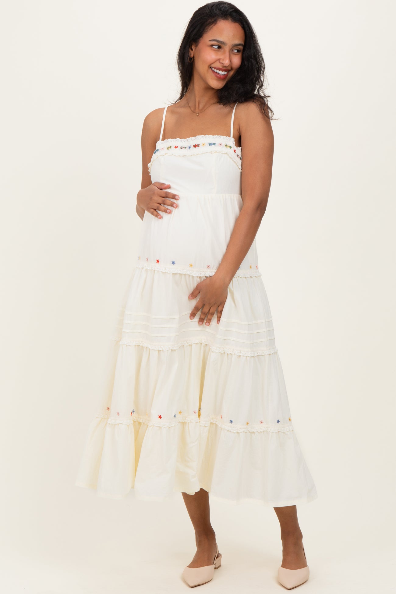 woman wearing a white tiered maternity dress with colorful embroidery and holding her baby bump