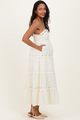 woman wearing a cream colored maternity dress with floral embroidery, tiered ruffles, and pockets, showing baby bump