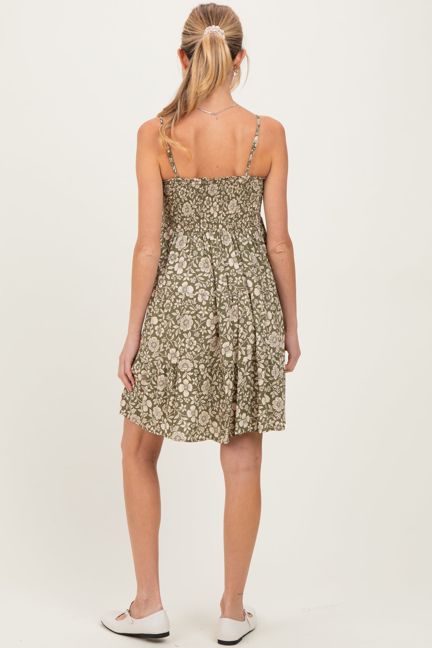 woman wearing a green floral maternity dress with adjustable straps and a relaxed fit, standing with her back to the camera