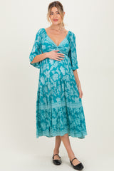 woman wearing a teal floral maternity dress with a deep v-neck and flowing sleeves, holding her baby bump