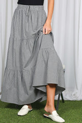 woman wearing a black and white gingham tiered maxi skirt and white slide sandals, holding the skirt hem