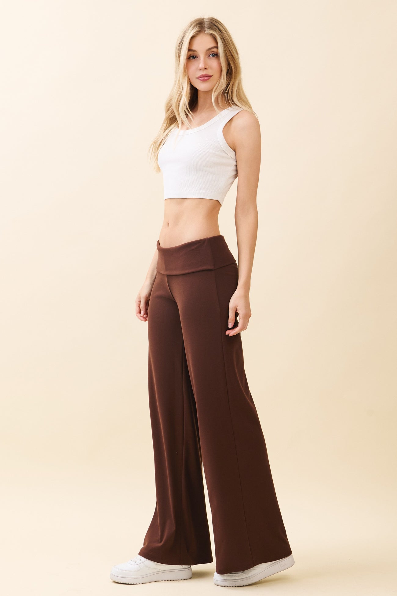 woman wearing a white crop top and brown wide leg pants, standing against a beige background