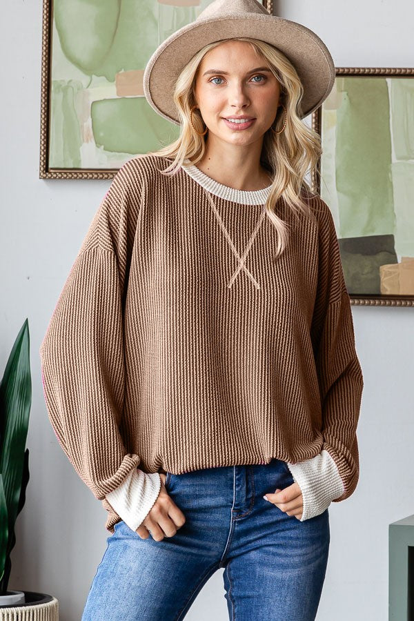 woman wearing a brown ribbed oversized sweater with white cuffs and a wide-brimmed hat, standing in a stylish interior