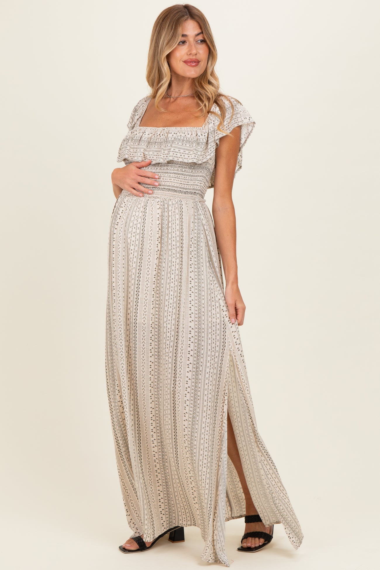 woman wearing a beige maternity maxi dress with ruffled sleeves and a front slit while holding her baby bump