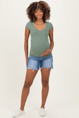 woman wearing a green ribbed maternity top and distressed denim shorts, holding her baby bump