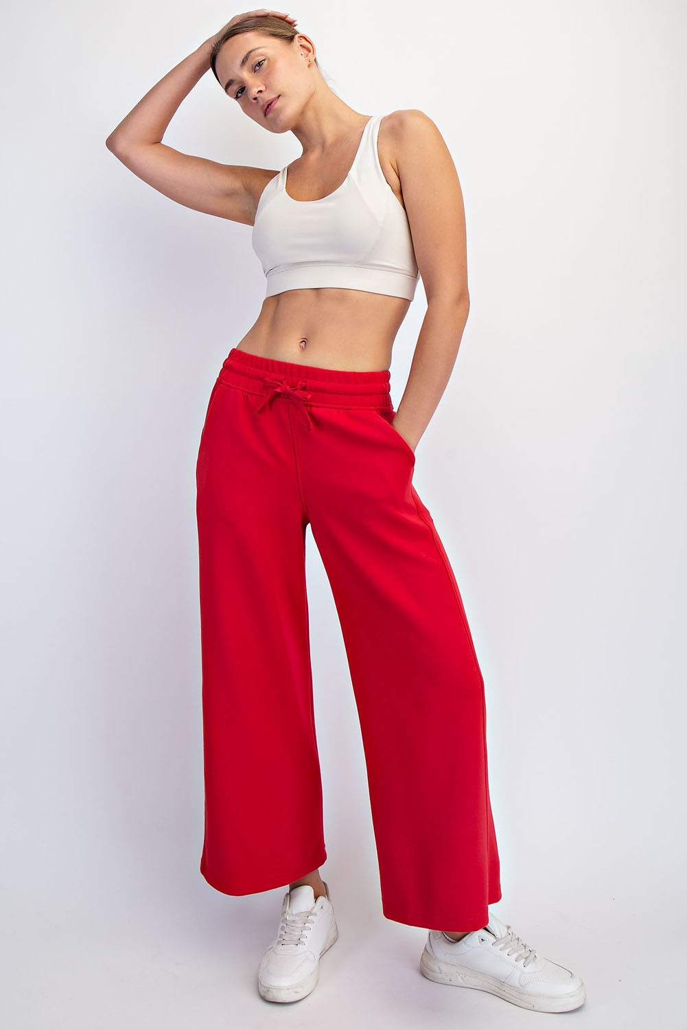 woman wearing a white crop top and red wide-leg sweatpants, posing with one hand on her hip and wearing white sneakers
