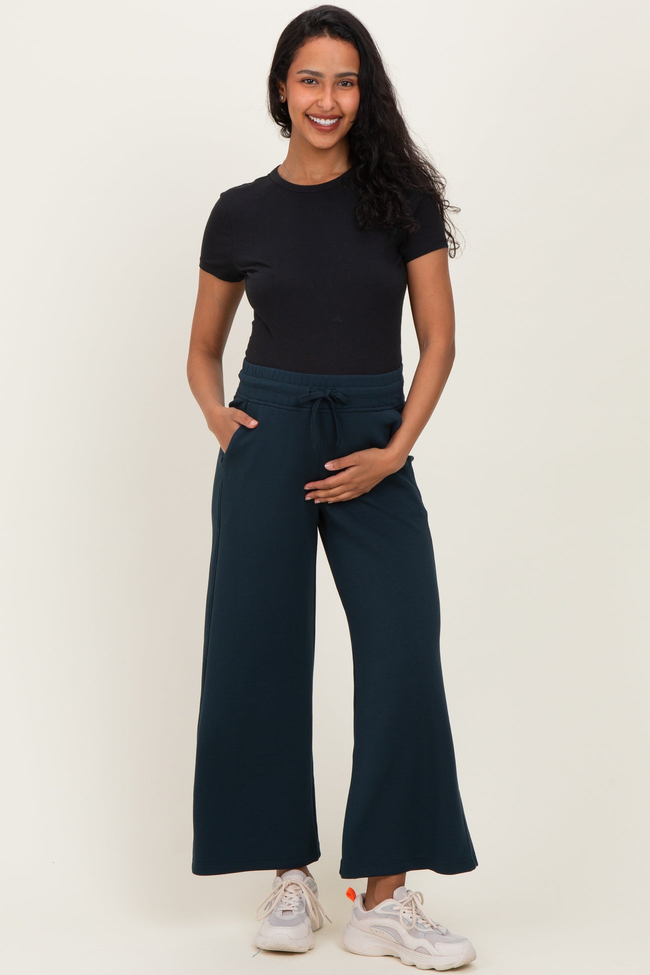 woman wearing a black short sleeve top and dark teal maternity wide-leg pants, holding her baby bump with one hand