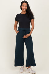 woman wearing a black short sleeve top and dark teal maternity wide-leg pants, holding her baby bump with one hand