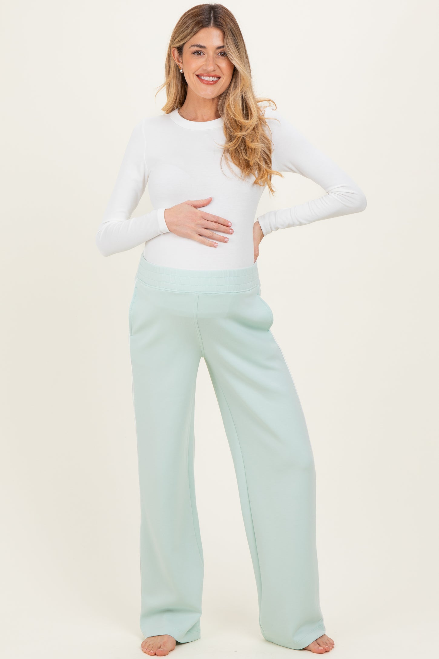 woman wearing a white long sleeve maternity top and light green maternity pants, holding her baby bump