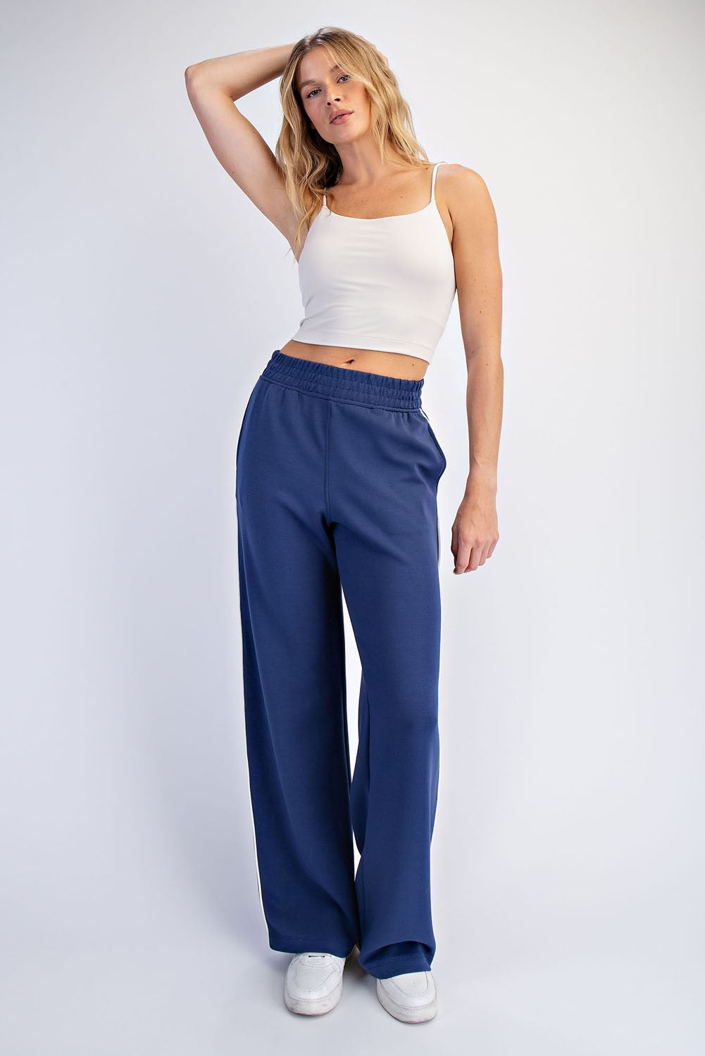 woman wearing a white spaghetti strap crop top and loose navy blue sweatpants, standing with one hand on her hip