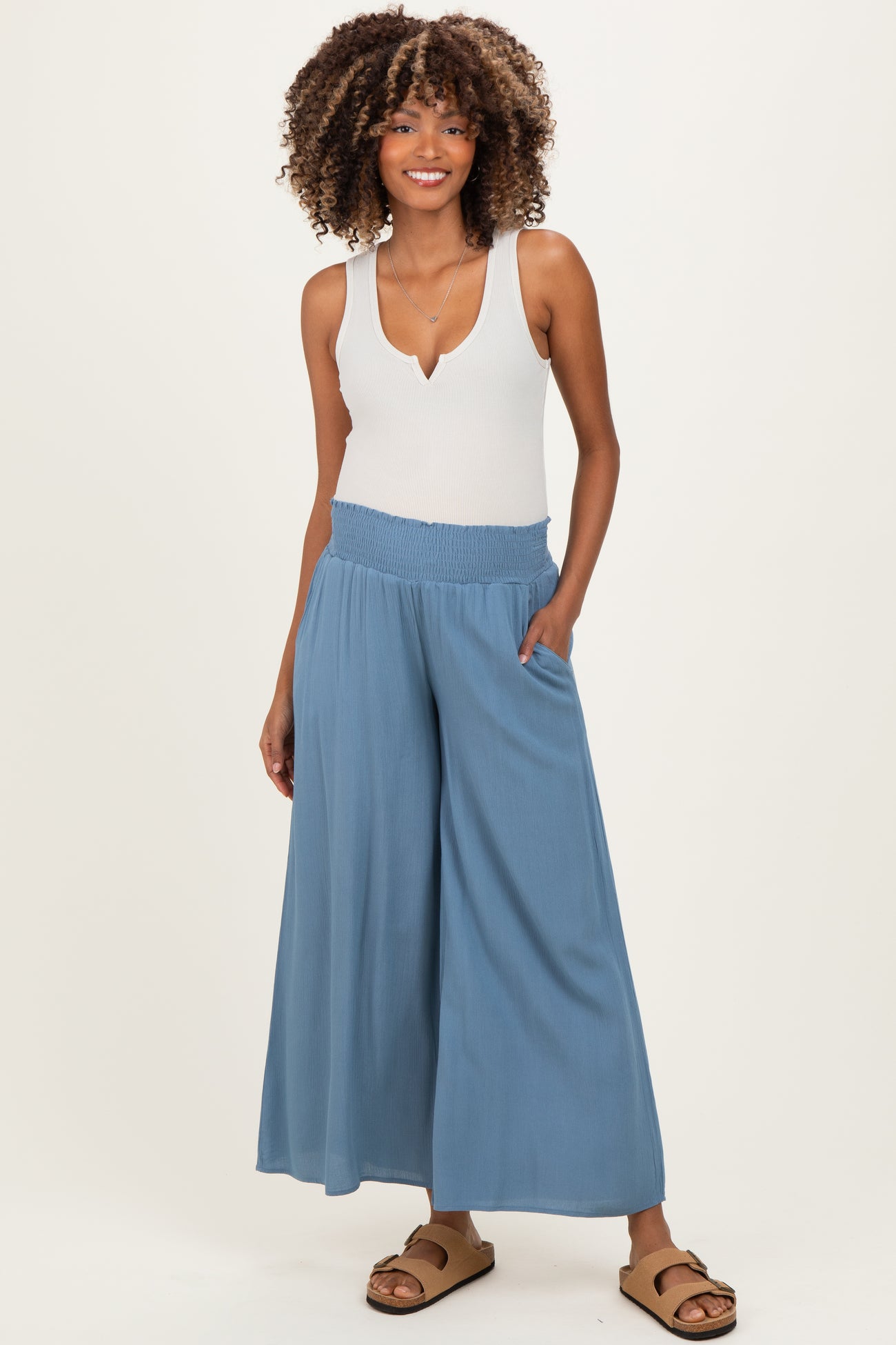 woman wearing a light blue maternity jumpsuit with wide legs and a smocked waistband, standing with hands in pockets