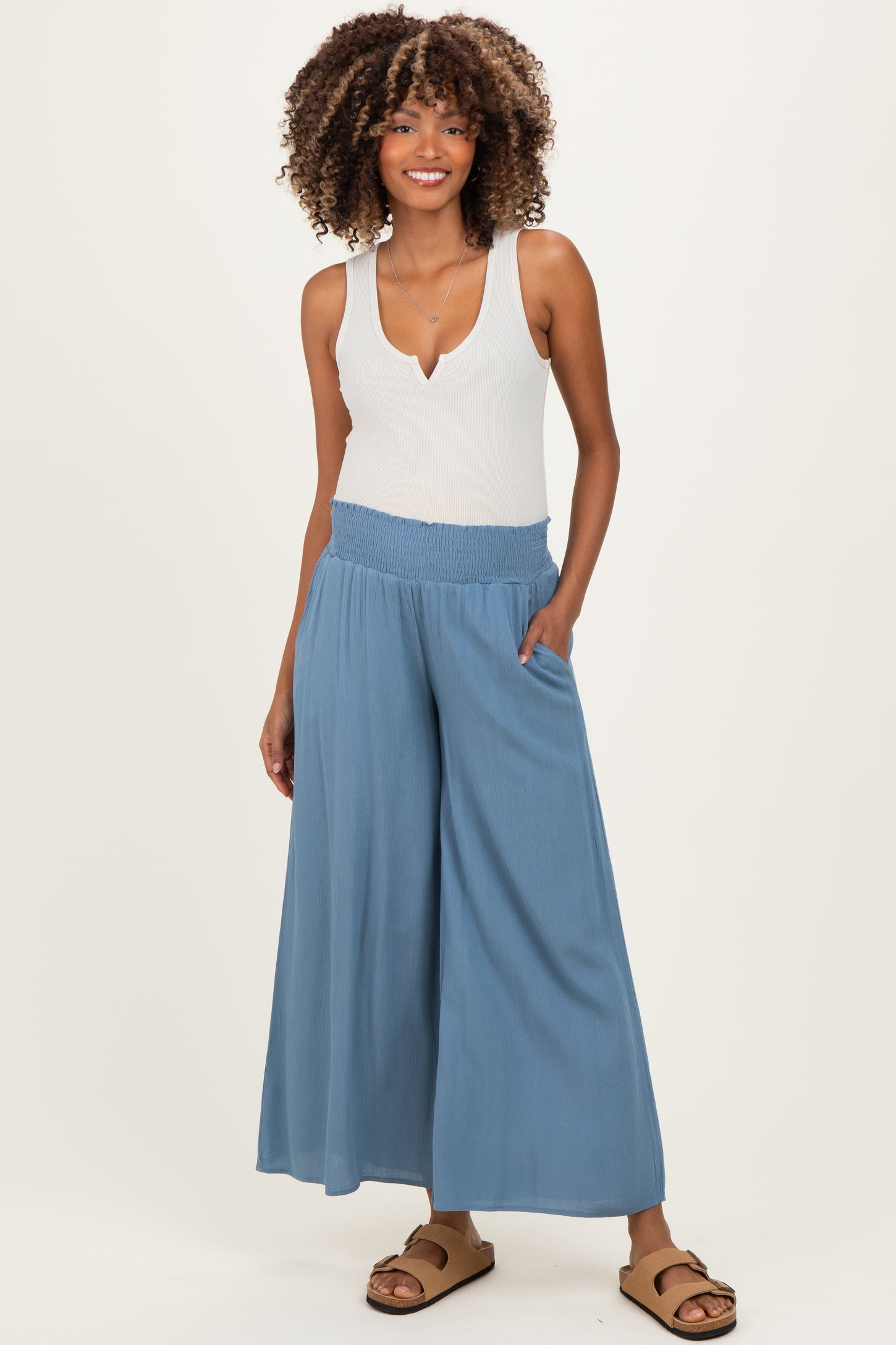 woman wearing a light blue maternity jumpsuit with wide legs and a smocked waistband, standing with hands in pockets