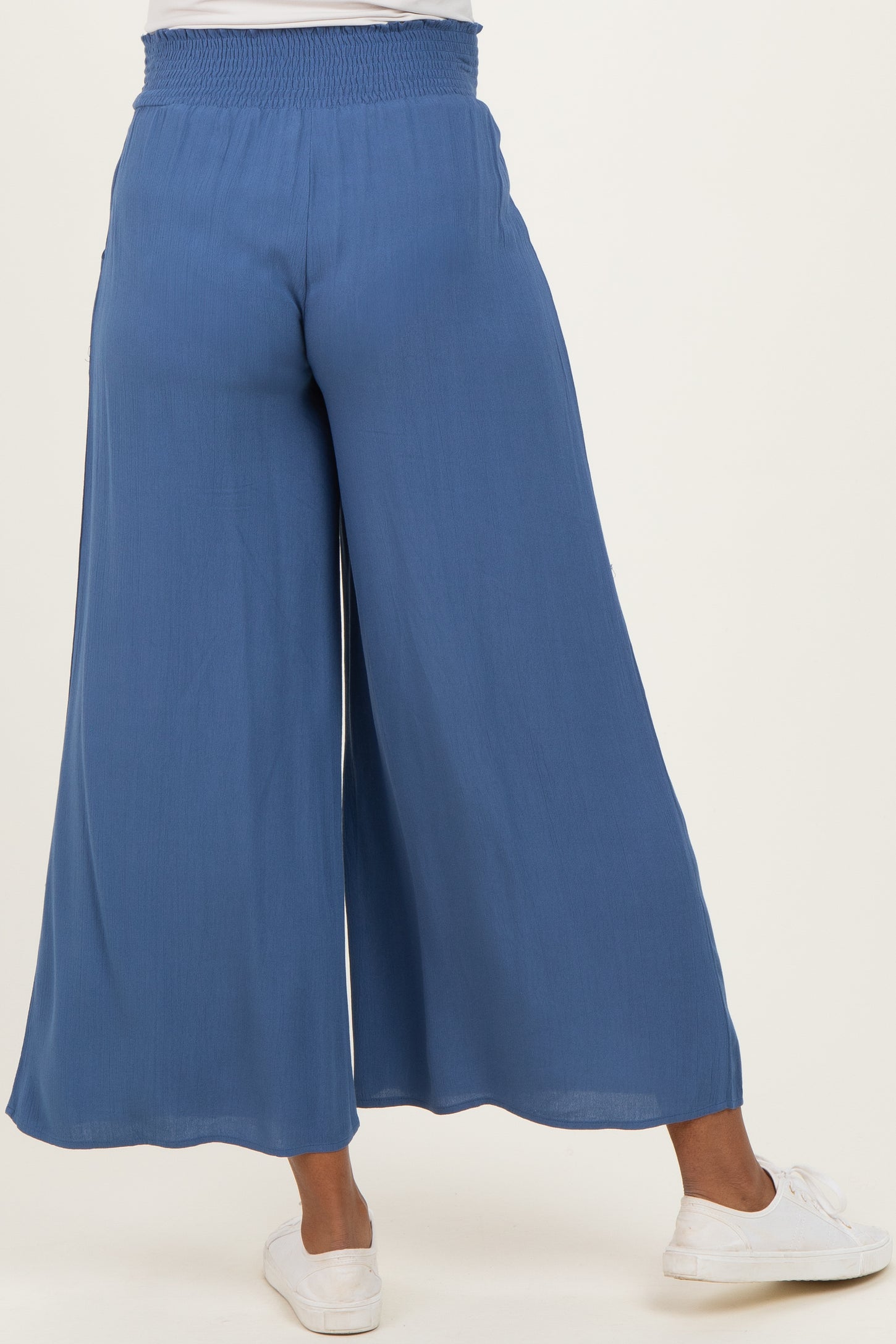 woman wearing blue maternity wide-leg pants, showing the back view with a smocked waistband and paired with white sneakers