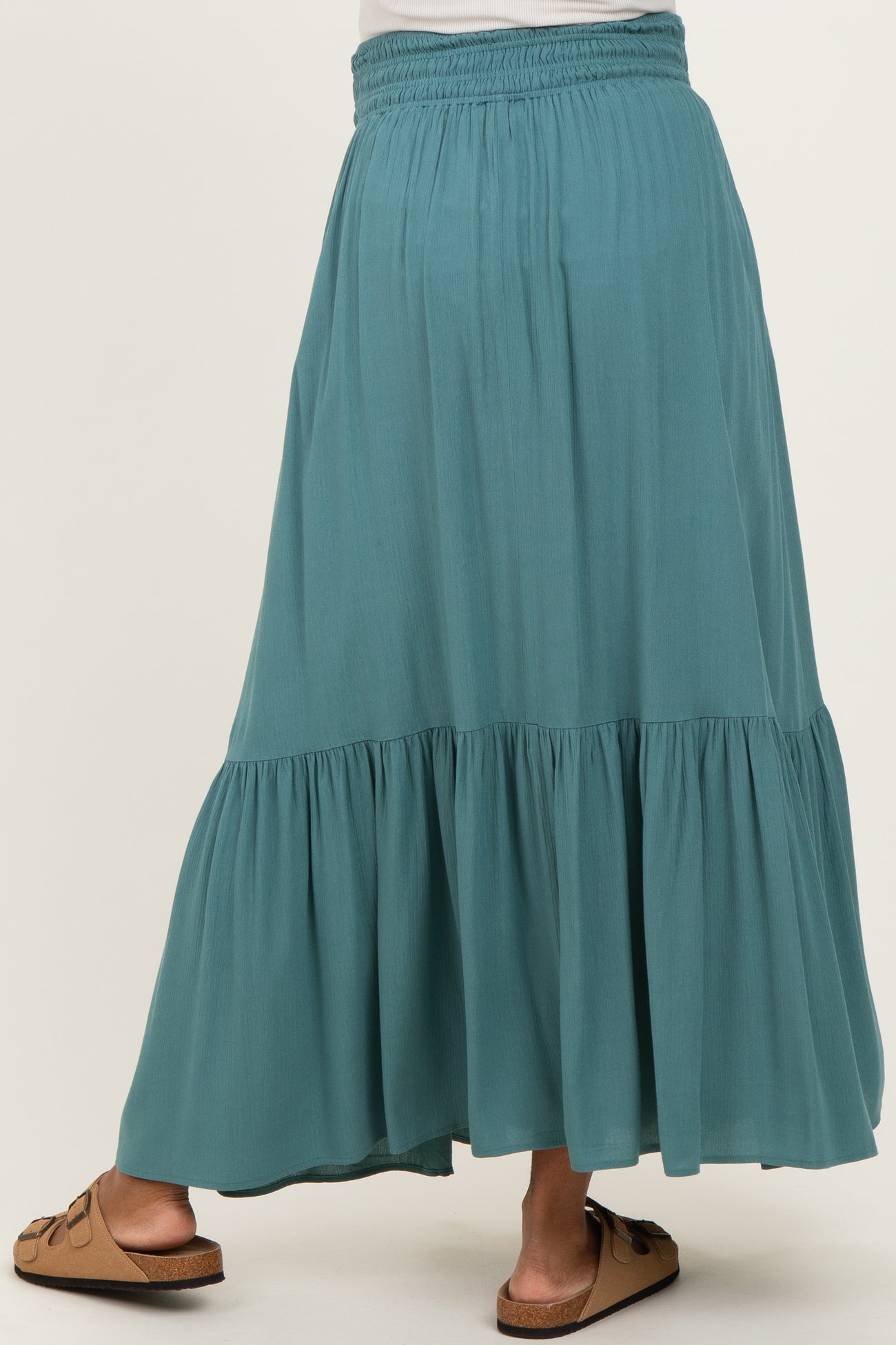 woman wearing a teal maternity maxi skirt with a tiered design and elastic waistband, standing with her back to the camera