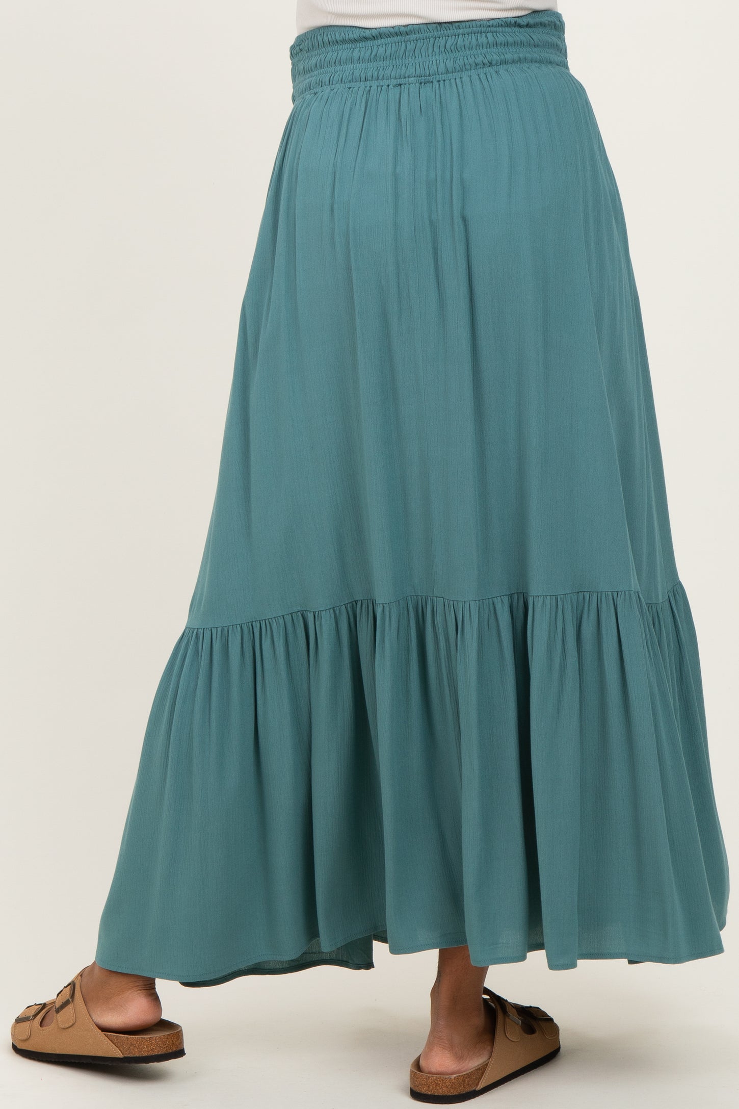 woman wearing a teal maternity maxi skirt with a tiered design and elastic waistband, standing with her back to the camera