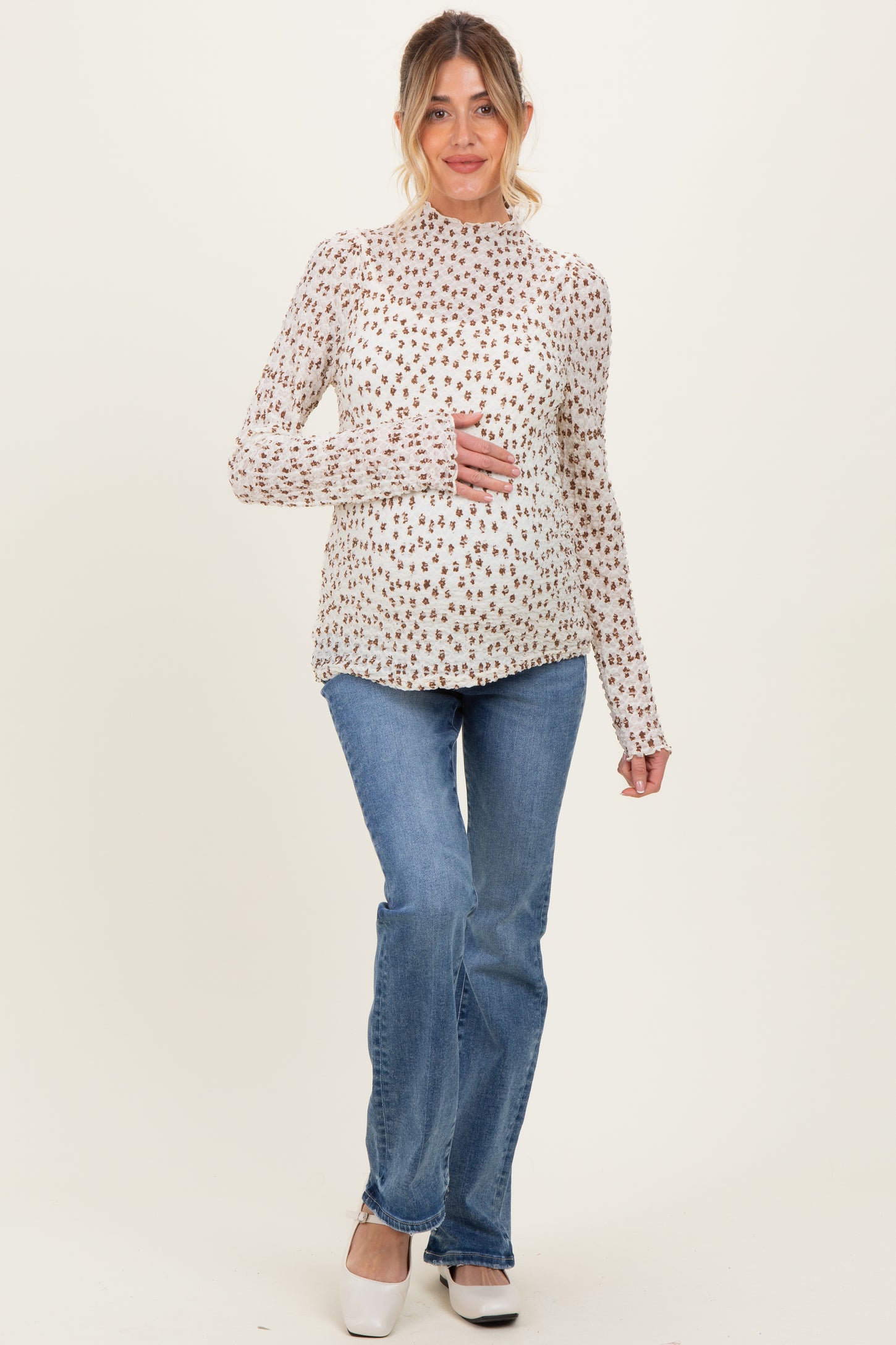 woman wearing a cream long sleeve maternity top with floral print and holding her baby bump, paired with blue flared jeans