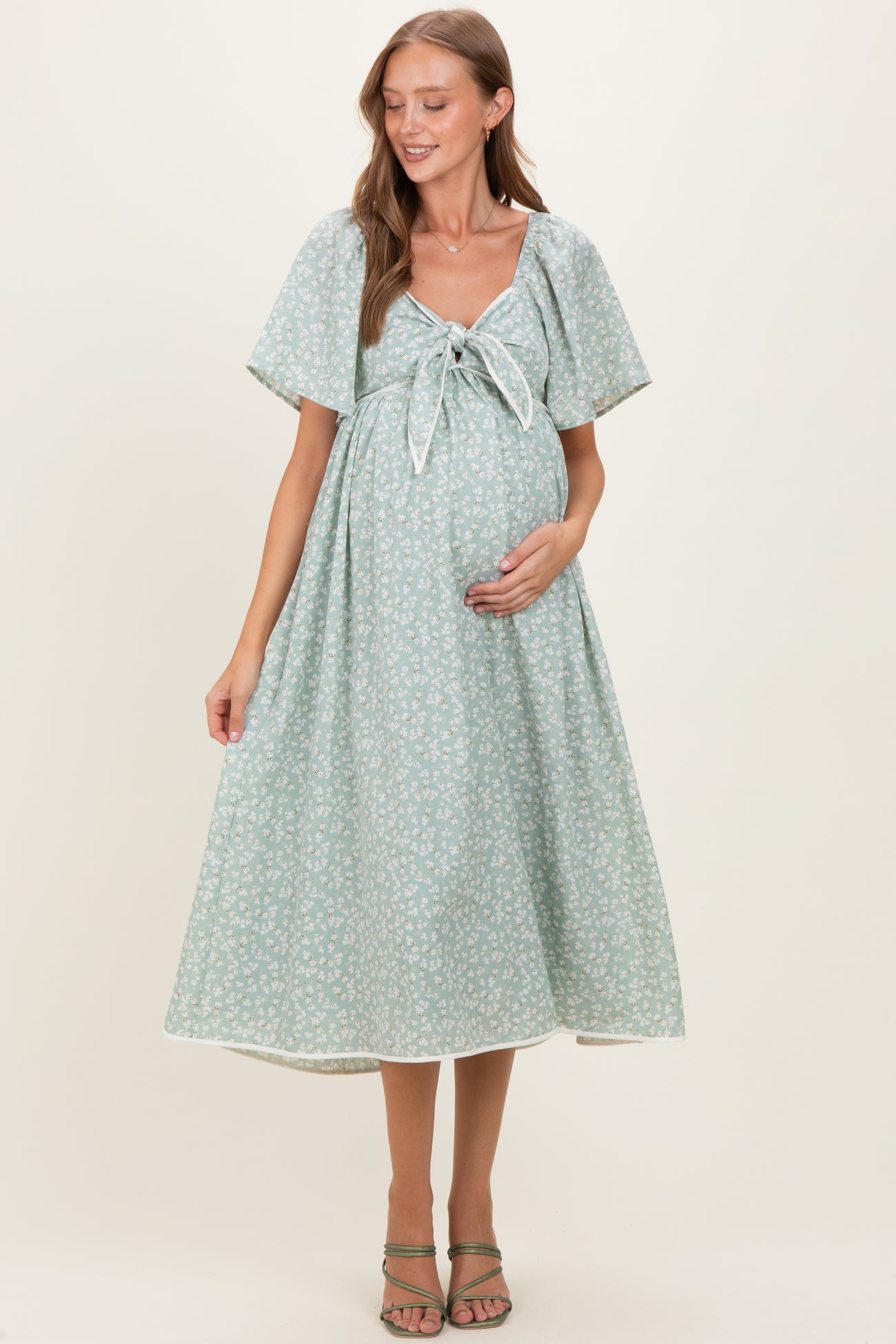 woman wearing a light green floral print maternity dress with a tie front, holding her baby bump