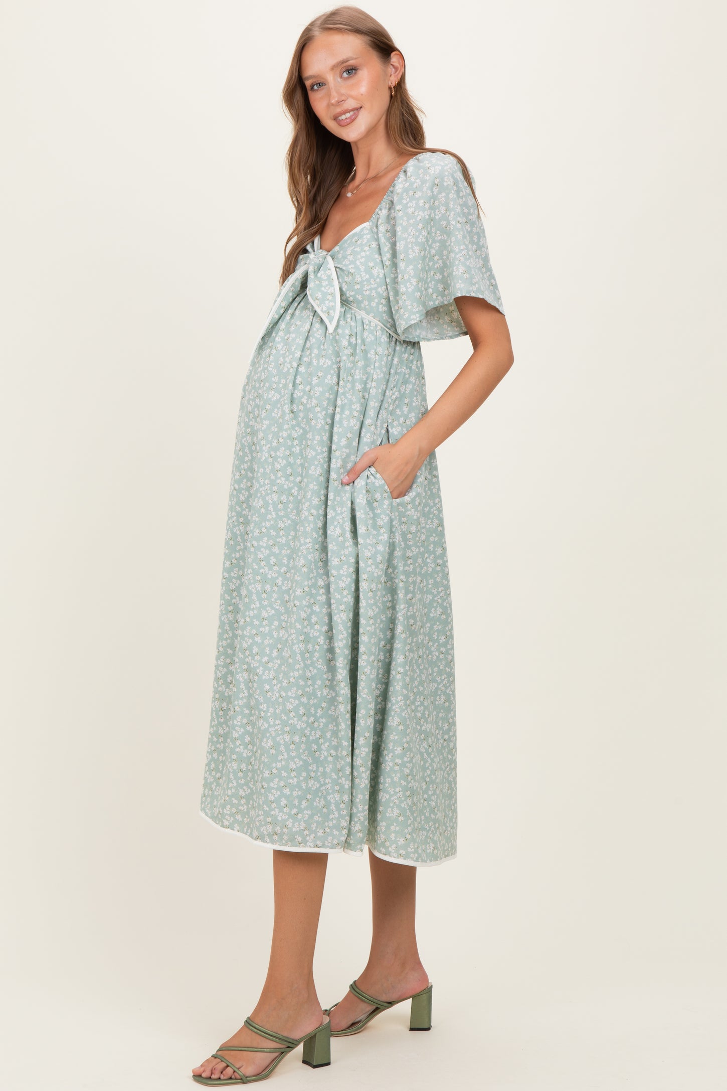 woman wearing a green floral print maternity dress with a bow detail and pockets, showing her baby bump