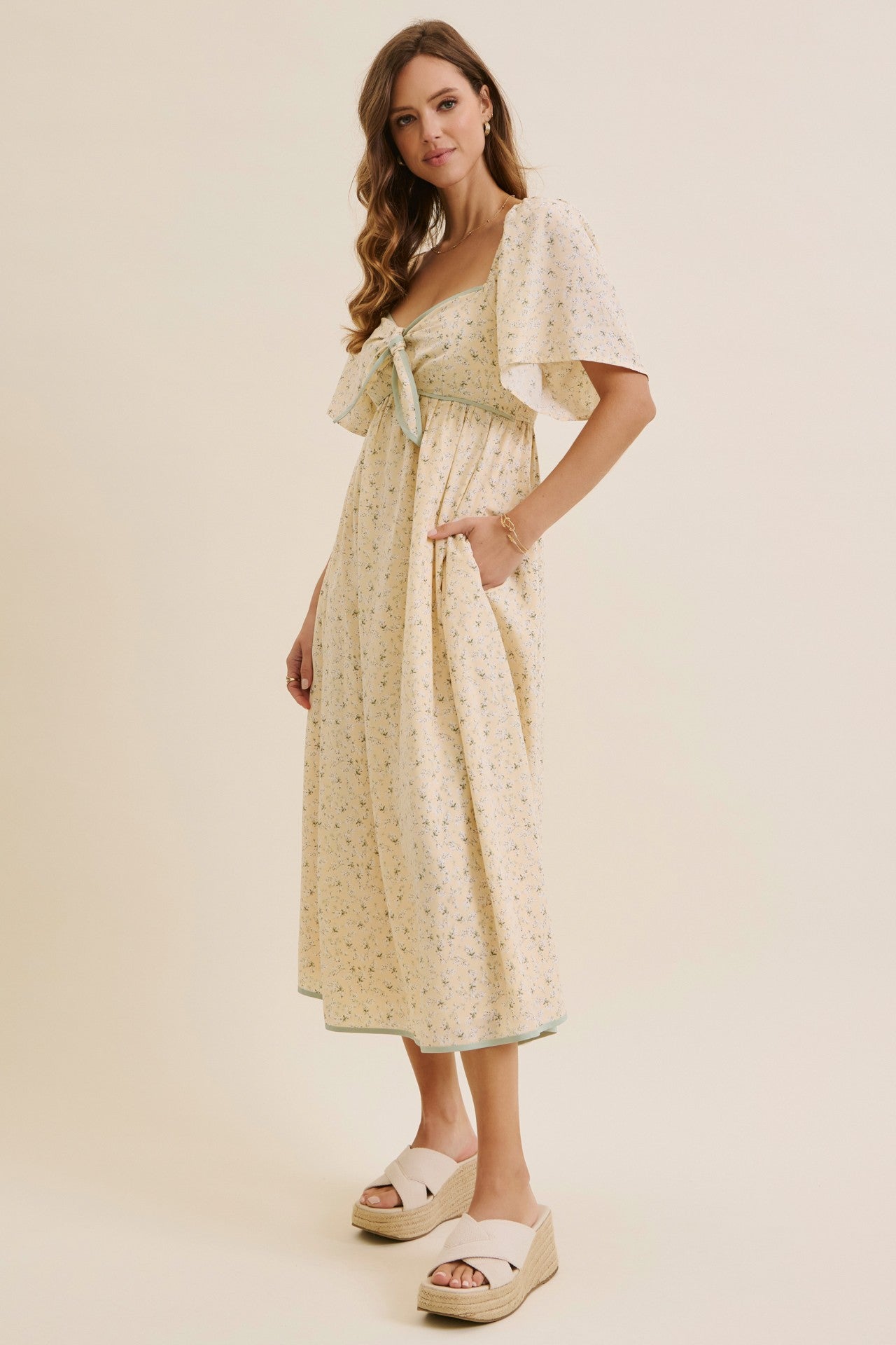 woman wearing a floral print yellow maxi dress with a tie-front detail and flutter sleeves, paired with white wedge sandals
