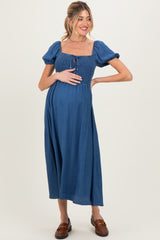 woman wearing a blue maternity dress with puff sleeves and a square neckline, gently holding her baby bump