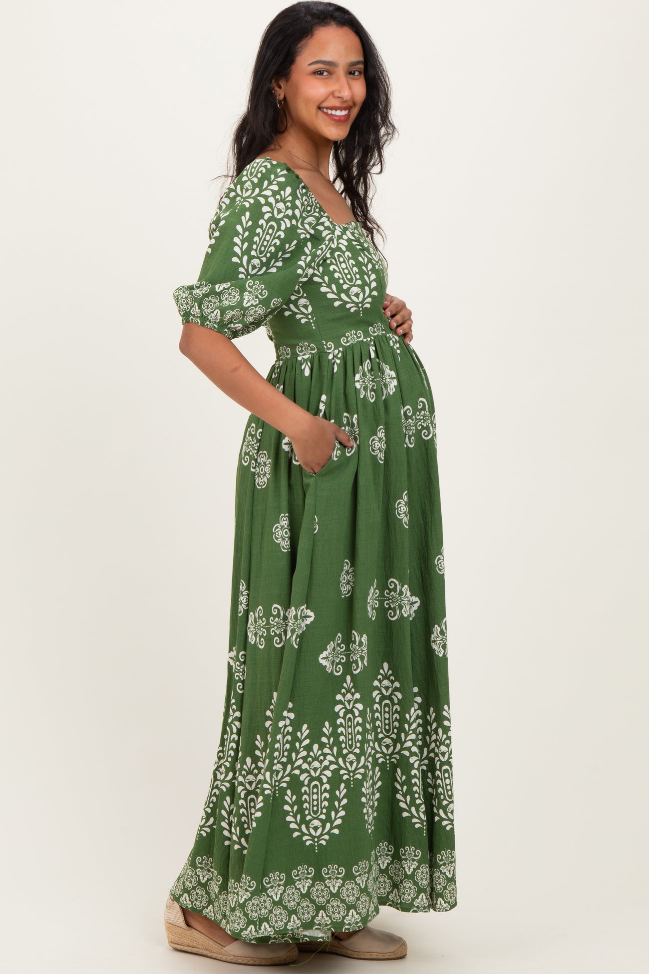 woman wearing a green maternity maxi dress with a white floral print and side pockets, showing her baby bump